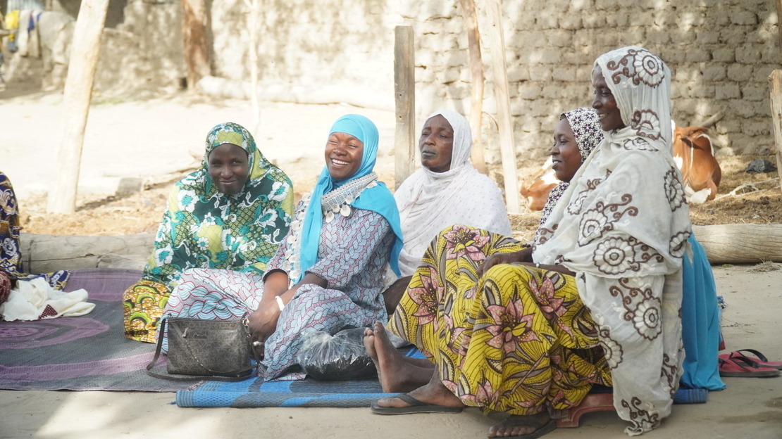 Five women in a semi-circle sitting on a mat smile and laugh. 