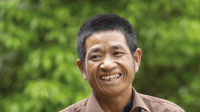 Portrait of Mr. Khamphong, a middle-aged man smiling wide, infront of a green nature background