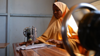 A young woman wearing a yellow veil sits at a table, sewing on her machine.