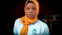Portrait of Saly: she's wearing an orange scarf and a T-shirt with the HI logo. In the background, is her sewing room.
