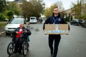 A man carrying a cardboard box walks down the street next to a man using a wheelchair.