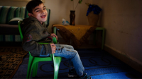 A young boy with cerebral palsy wearing jeans and a hooded sweatshirt sits in a green plastic chair in his living room.