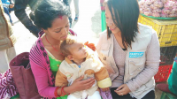 A mother holds her toddler son with clubfoot in her lap while a female HI technician smiles 